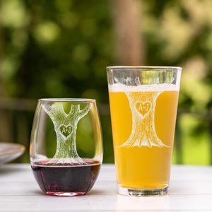 Puede incluir: Dos vasos personalizados con un dise&ntilde;o de &aacute;rbol grabado en la parte delantera. El &aacute;rbol tiene un coraz&oacute;n en el centro con las iniciales "A+J" dentro. Un vaso est&aacute; lleno de vino tinto y el otro de una bebida amarilla clara.