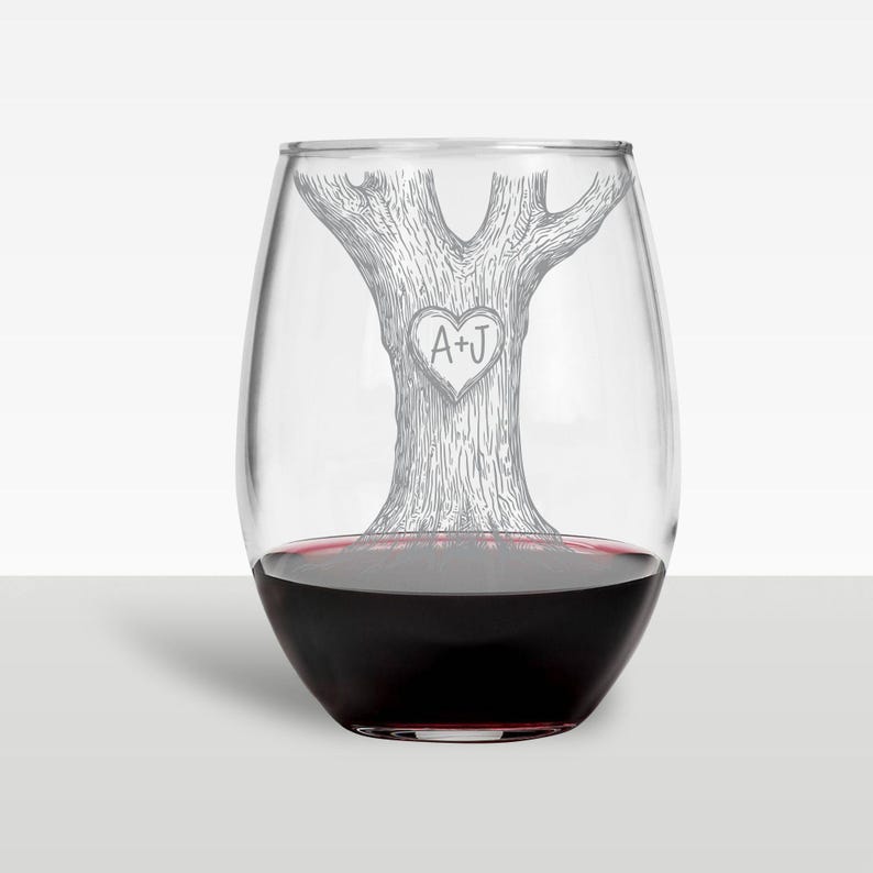 Puede incluir: Una copa de vino sin tallo transparente con un dise&ntilde;o de &aacute;rbol grabado en el lateral. El &aacute;rbol tiene un coraz&oacute;n tallado con las iniciales "A+J" en su interior.