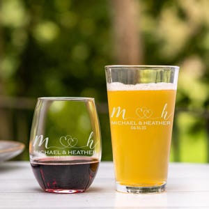 May include: Two personalized glasses, one for wine and one for beer, with the initials "M" and "H" intertwined with hearts and the text "Michael & Heather 04.16.25".