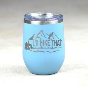 May include: A light blue stainless steel tumbler with a white lid and a silver inscription that reads "I'd Hike That". The inscription is accompanied by a graphic of mountains and trees.