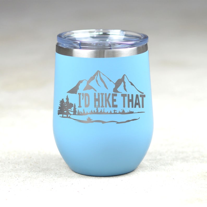 May include: A light blue stainless steel tumbler with a white lid and a silver inscription that reads "I'd Hike That". The inscription is accompanied by a graphic of mountains and trees.