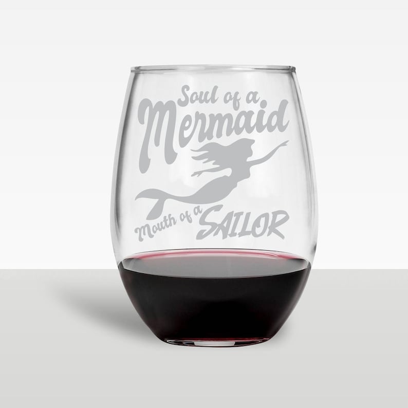 May include: A stemless wine glass filled with red wine. The glass has a gray mermaid graphic and the text "Soul of a Mermaid, Mouth of a Sailor". The glass is clear and sits on a white surface.