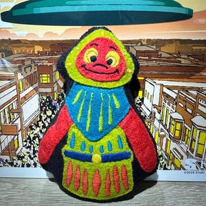 May include: A felted plush figure featuring a red face with yellow eyes, a blue chest piece with yellow accents, and a lime green body with red and blue detailing. The figure has a black outline and is shaped like a stylized character with a hood.