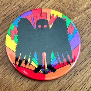 May include: Round button with a black silhouette of a winged creature with red eyes, set against a rainbow background. The word "THW" is in the upper portion of the button. The design is bold and colorful.