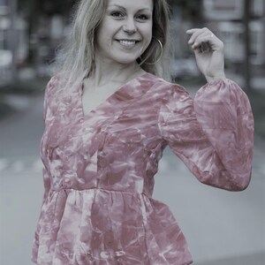 May include: A pink and white patterned blouse with a V-neck and long, puffy sleeves. The blouse has a peplum waist and is paired with white pants. The woman is smiling and wearing large hoop earrings.