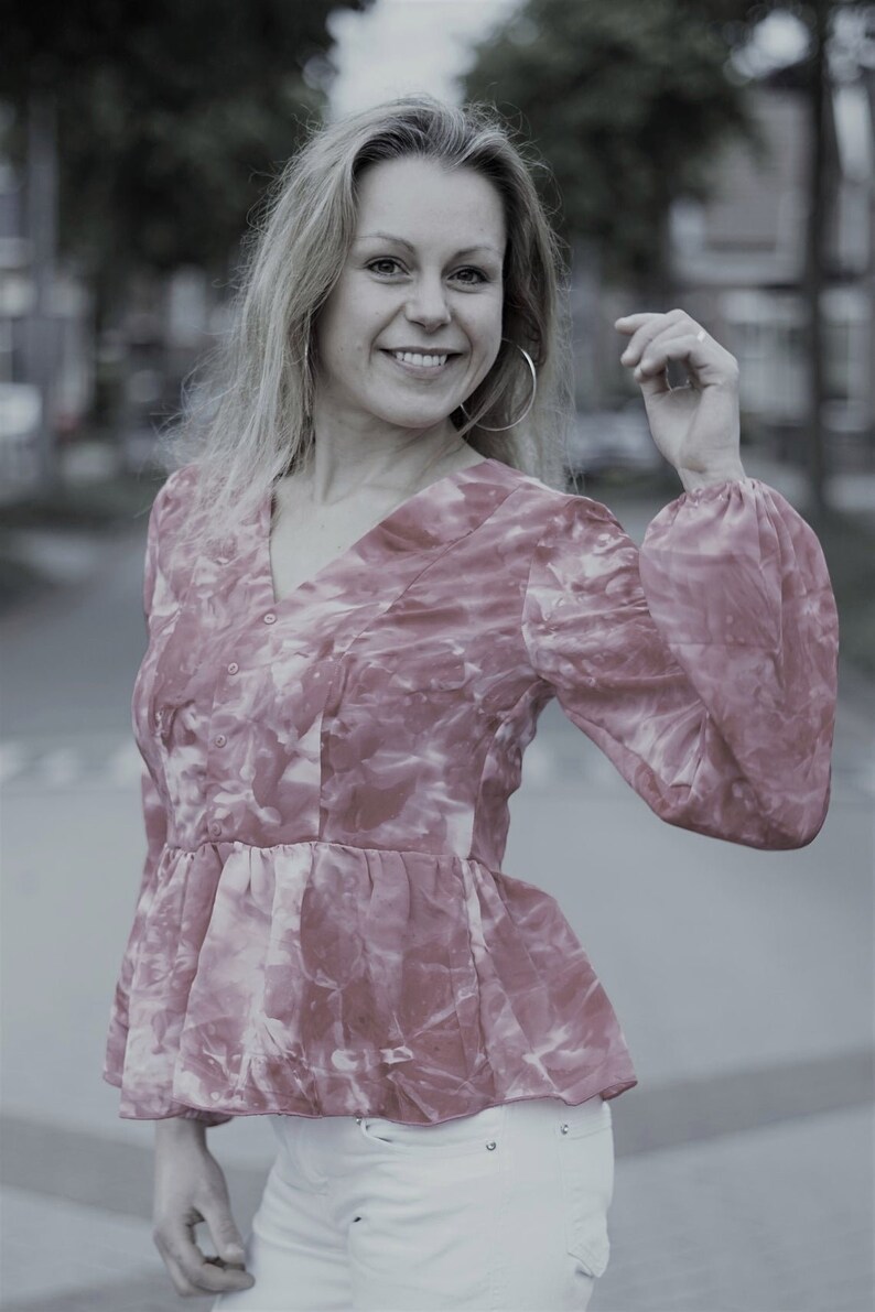 May include: A pink and white patterned blouse with a V-neck and long, puffy sleeves. The blouse has a peplum waist and is paired with white pants. The woman is smiling and wearing large hoop earrings.