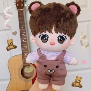 May include: A plush doll with brown hair and bear ears, wearing a striped shirt and brown overalls with a bear face. The doll has large, expressive eyes and a small pink nose. A guitar is in the background.