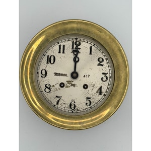 May include: A round wall clock featuring a wide, polished brass frame and a weathered white face. The dial displays black Arabic numerals from one to twelve, two black hands, and two small winding holes. The surface shows visible wear.