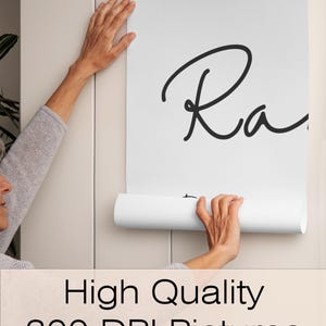 May include: A white poster with the word "Ra" in black script is being held up against a light beige wall. The poster is unrolled, revealing the text. The bottom of the image has the text "High Quality 300 DPI Pictures".