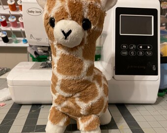 Könnte beinhalten: Ein Plüsch-Giraffe-Spielzeug mit braunen und weißen Flecken. Die Giraffe hat ein weißes Gesicht mit schwarzen Augen und einer schwarzen Nase. Die Giraffe steht auf einer grauen Oberfläche.