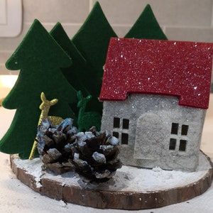 May include: A festive miniature Christmas scene. It features a silver glitter house with a red roof, green felt trees, and gold deer figurines. Two pine cones with blue glitter sit on a wooden base, creating a wintery display.