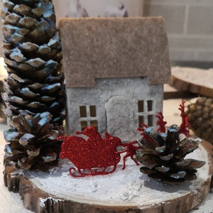 May include: A Christmas-themed miniature scene. It features a small house with a brown roof, snow, pine cones, and a red glitter sleigh with reindeer. The scene is set on a wooden slice, creating a winter wonderland.