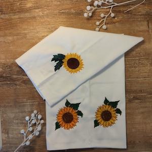 Puede incluir: Toallas de cocina blancas con bordados de girasoles. Los girasoles tienen pétalos amarillos, centros marrones y hojas verdes. Las toallas están dobladas sobre una superficie de madera.