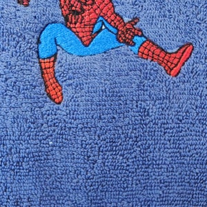 May include: Blue towel with an embroidered Spider-Man design in red, blue, and black. The name "MATTIA" is embroidered in red at the bottom. The towel has a textured surface.