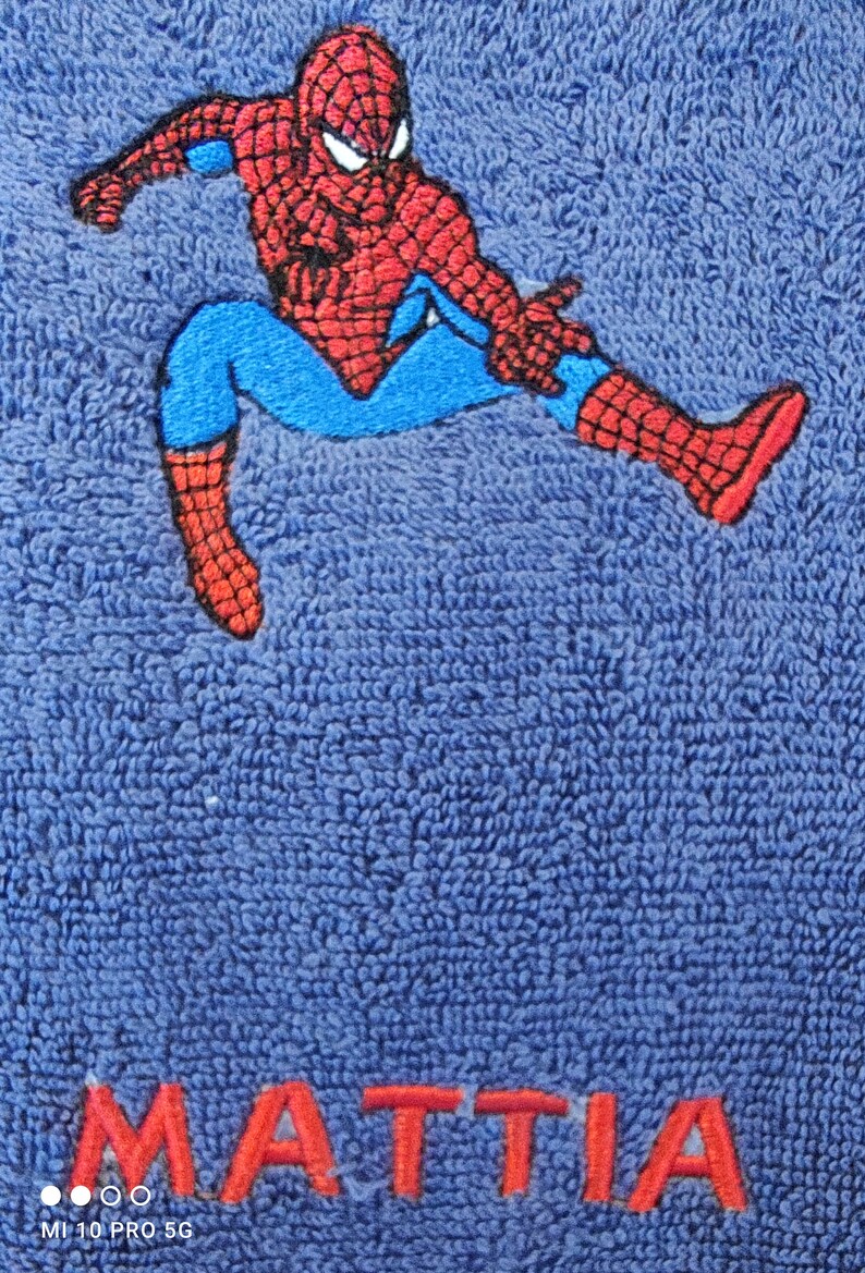 May include: Blue towel with an embroidered Spider-Man design in red, blue, and black. The name "MATTIA" is embroidered in red at the bottom. The towel has a textured surface.