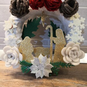 May include: A festive Christmas wreath with a nativity scene design. The wreath is decorated with felt flowers, glitter, and a star. The scene depicts Mary, Joseph, and baby Jesus, finished with gold glitter. The wreath is decorated with red, white, and brown felt flowers.