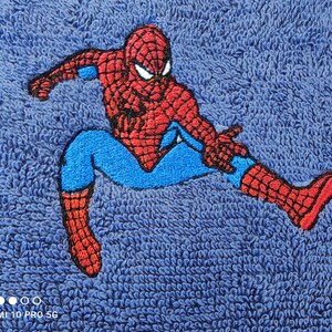 May include: Embroidered Spider-Man patch in red, blue, and black on a blue textured fabric. The superhero is shown in a dynamic pose. The patch is likely for clothing or accessories. The fabric has a textured appearance.