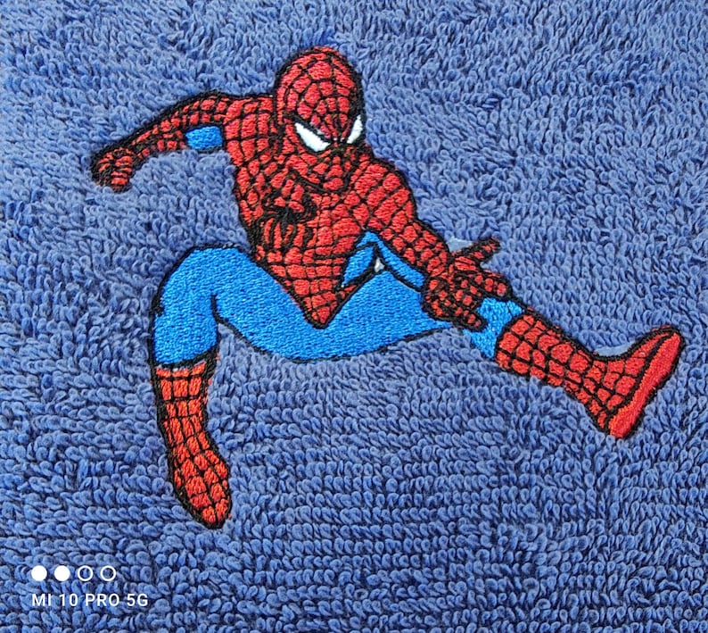 May include: Embroidered Spider-Man patch in red, blue, and black on a blue textured fabric. The superhero is shown in a dynamic pose. The patch is likely for clothing or accessories. The fabric has a textured appearance.