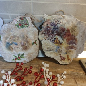 May include: Two rustic wooden Christmas ornaments. One depicts a nativity scene with holly, the other a winter landscape with a cabin and deer. Twine loops for hanging. Red and white berry sprigs are in the foreground.