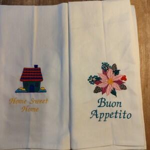 Puede incluir: Dos paños de cocina blancos con diseños bordados. Uno presenta una casa con el texto "Home Sweet Home" en amarillo. El otro tiene un diseño floral con el texto "Buon Appetito" en turquesa.