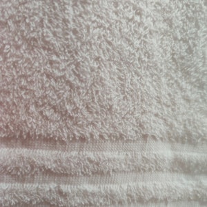 May include: Close-up of a white, fluffy towel. The terry cloth texture is visible, with a pattern of horizontal stripes near the bottom. The towel appears soft and absorbent, ideal for bathroom use.