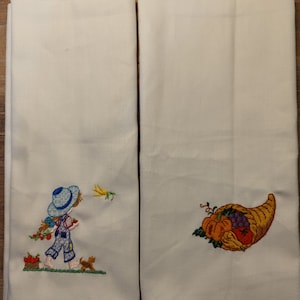 Puede incluir: Dos paños de cocina blancos bordados. Uno presenta a un niño con peto y sombrero, llevando una cesta de manzanas rojas, con un perro pequeño. La otra toalla tiene un diseño de cuerno de la abundancia con varias frutas y verduras.