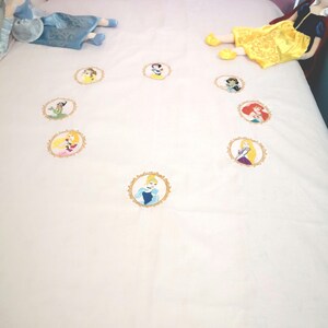 May include: White bedspread featuring embroidered portraits of Disney princesses in gold frames. Plush dolls of Cinderella, Snow White, and another princess are on the bed. The bed has a purple headboard.