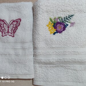 May include: Two white bath towels with embroidered designs. One towel features a magenta butterfly, the other a floral arrangement with yellow, purple, and green accents. Both towels have a textured surface and a decorative stripe.