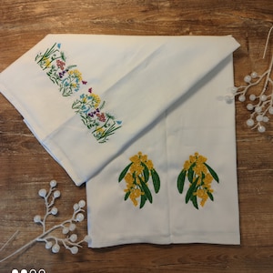 Puede incluir: Toalla de cocina de algodón blanco con diseños florales bordados. Un borde presenta un borde colorido de flores silvestres, mientras que el otro tiene dos racimos de flores de mimosa amarillas con hojas verdes. La toalla está doblada sobre una superficie de madera.