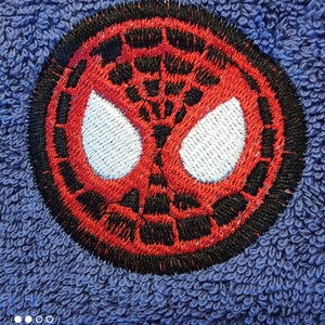 May include: Circular embroidered patch of the Spider-Man mask design. The patch features a red and black web pattern with white eye shapes, set against a blue, textured background. The design is detailed.