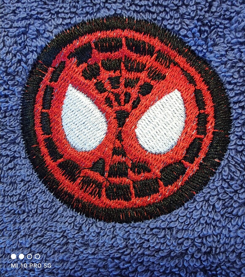 May include: Circular embroidered patch of the Spider-Man mask design. The patch features a red and black web pattern with white eye shapes, set against a blue, textured background. The design is detailed.