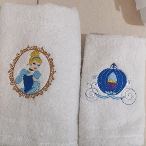 Peut inclure: Deux serviettes blanches brodées. L'une présente Cendrillon dans un cadre doré, vêtue d'une robe bleue. L'autre serviette a un motif de carrosse bleu. Les deux serviettes sont en matière douce et absorbante.