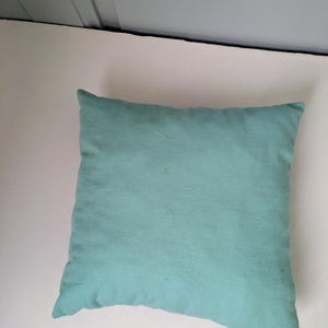 K&ouml;nnte beinhalten: Ein quadratisches, helles tealfarbenes Kissen. Das Kissen hat ein weiches, strukturiertes Aussehen und ist wahrscheinlich aus Stoff. Das Kissen ist einfarbig und scheint neu zu sein. Das Kissen ist ein dekorativer Artikel.