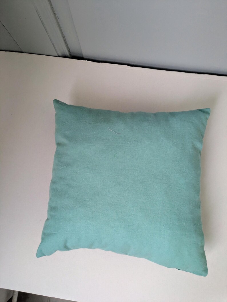 K&ouml;nnte beinhalten: Ein quadratisches, helles tealfarbenes Kissen. Das Kissen hat ein weiches, strukturiertes Aussehen und ist wahrscheinlich aus Stoff. Das Kissen ist einfarbig und scheint neu zu sein. Das Kissen ist ein dekorativer Artikel.