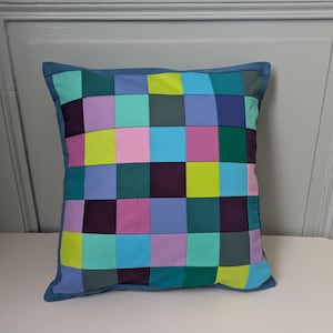 Peut inclure: Coussin carré décoratif avec un motif patchwork. Le coussin présente un motif quadrillé de carrés colorés dans les tons de bleu, vert, violet, rose et jaune. Le coussin a une bordure bleue unie.