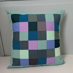 Peut inclure: Coussin carré décoratif avec un motif patchwork. Le coussin présente un motif quadrillé de différentes nuances de bleu, vert, violet et gris. La bordure est de couleur vert clair uni. Le coussin est de forme carrée.