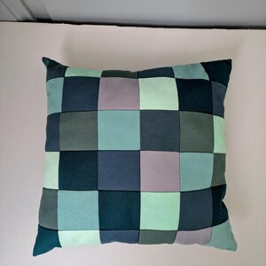 K&ouml;nnte beinhalten: Quadratisches Kissen mit Patchwork-Design. Das Kissen hat ein Rastermuster aus verschiedenen Blau-, Gr&uuml;n- und Graut&ouml;nen. Das Kissen ist ein dekoratives Element f&uuml;r ein Bett oder Sofa.