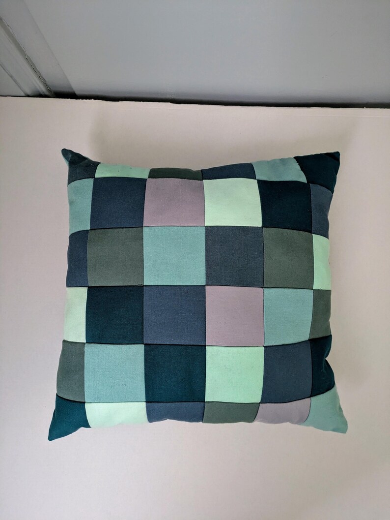 K&ouml;nnte beinhalten: Quadratisches Kissen mit Patchwork-Design. Das Kissen hat ein Rastermuster aus verschiedenen Blau-, Gr&uuml;n- und Graut&ouml;nen. Das Kissen ist ein dekoratives Element f&uuml;r ein Bett oder Sofa.
