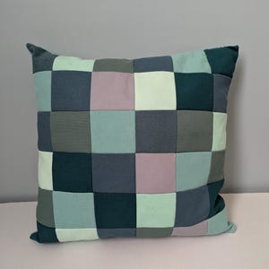 K&ouml;nnte beinhalten: Quadratisches Patchwork-Kissen in T&uuml;rkis-, Grau- und Lavendelt&ouml;nen. Das Kissen weist ein Rastermuster aus Stoffquadraten auf, das ein geometrisches Design ergibt. Es hat ein weiches, einladendes Aussehen, passend f&uuml;r die Inneneinrichtung.