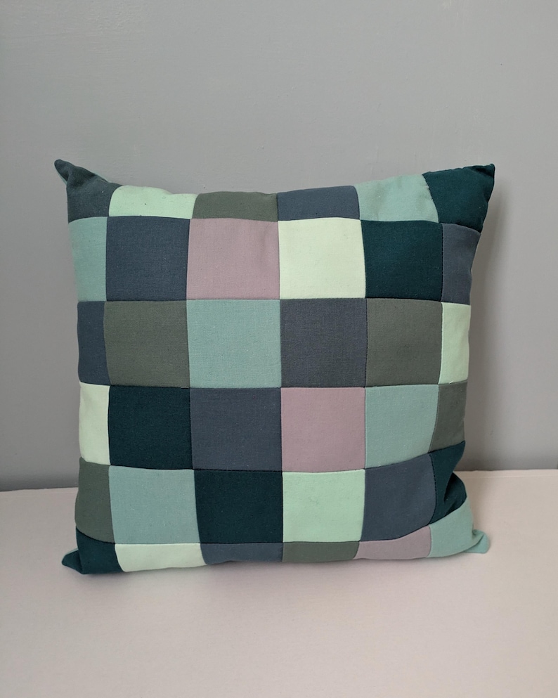 K&ouml;nnte beinhalten: Quadratisches Patchwork-Kissen in T&uuml;rkis-, Grau- und Lavendelt&ouml;nen. Das Kissen weist ein Rastermuster aus Stoffquadraten auf, das ein geometrisches Design ergibt. Es hat ein weiches, einladendes Aussehen, passend f&uuml;r die Inneneinrichtung.