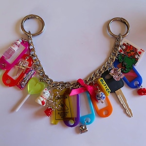 May include: A silver chain keychain with various charms, including a miniature water bottle, a cross, a lollipop, a lucky cat, and a playing card. Other charms include a key, a heart, and a caution sign.