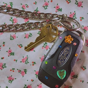Puede incluir: Una llave de coche negra con una cadena plateada y una llave con la palabra "KWI" en ella. La llave tiene diamantes de imitaci&oacute;n coloridos y un diamante de imitaci&oacute;n en forma de coraz&oacute;n. La llave est&aacute; sobre un fondo de tela floral.