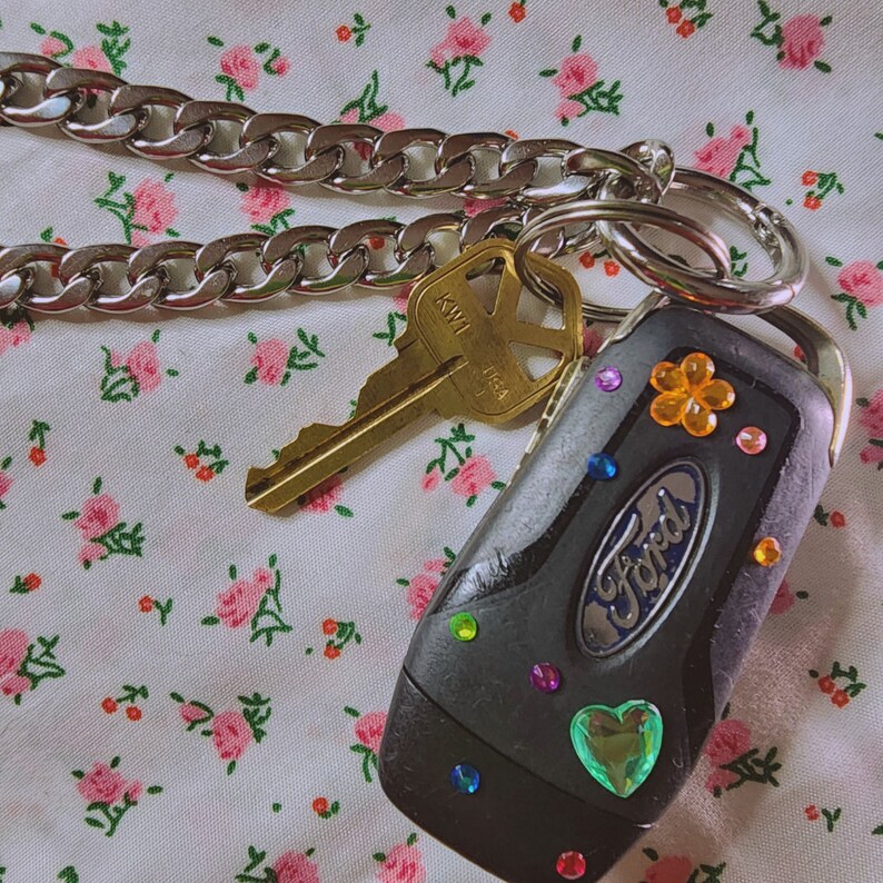 Puede incluir: Una llave de coche negra con una cadena plateada y una llave con la palabra "KWI" en ella. La llave tiene diamantes de imitaci&oacute;n coloridos y un diamante de imitaci&oacute;n en forma de coraz&oacute;n. La llave est&aacute; sobre un fondo de tela floral.