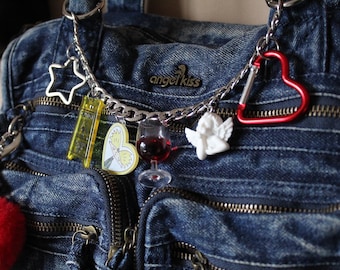May include: Close-up of a denim handbag with multiple charms. Charms include a star, a yellow ticket, a heart with a butterfly, a wine glass, a white angel, and a red heart-shaped carabiner. The bag has multiple zippers and the text "angel kiss".