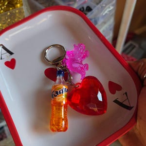 May include: A keychain with a miniature Fanta bottle, a red heart-shaped charm, and a pink dragon charm. The keychain is on a white tray with red trim and playing card symbols.