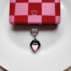 May include: A black and pink lips charm with a tongue sticking out, hanging from a silver chain. The charm is attached to a red and pink checkered phone case.