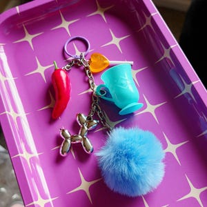 May include: A collection of keychains with various charms, including a red chili pepper, a silver balloon dog, a blue teacup, a heart-shaped lollipop, and a fluffy blue pom-pom. The keychains are arranged on a purple tray with a starburst pattern.