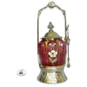 May include: An antique ruby red glass biscuit jar with a silver-toned metal handle and base. The jar features a floral design and a silver lid. The handle is adorned with a flower at the top. A spoon rests on the side.