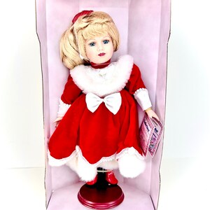 May include: A porcelain doll wearing a red velvet dress with white fur trim and a white bow. The doll has blonde hair and blue eyes. The doll is in a pink box with a white label that says "The Original Samantha".