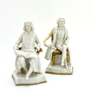May include: Two porcelain figurines of famous composers. The figurine on the left is seated and holding a scroll. The figurine on the right is seated on a chair and is labeled "Schubert".